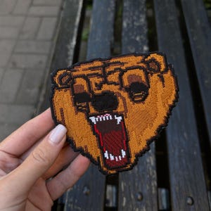 Peut inclure: Écusson brodé représentant une tête d'ours pixélisée avec des lunettes et une bouche ouverte. Le patch est principalement marron avec des détails noirs et beiges, et une bordure noire. Design rétro.