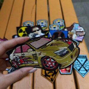 May include: Embroidered patch featuring a gold-colored sports car with red and pink accents. The patch has a detailed design with text and is surrounded by other patches with various designs.