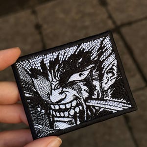 May include: A rectangular embroidered patch featuring a high-contrast black and white graphic illustration of a character with an intense expression, bared teeth, and spiky hair, finished with a thick black stitched border around the edges.