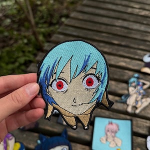 May include: An embroidered patch featuring a stylized anime character head with short, light blue hair, large eyes with bright red irises, and a tan-colored collar. The design is outlined in black thread and has a distinct, expressive facial expression.