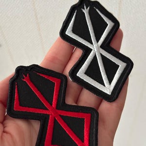 Puede incluir: Dos parches bordados. Uno es negro con un diseño geométrico blanco. El otro es negro con un diseño geométrico rojo. Ambos parches tienen un borde negro.