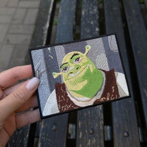 Peut inclure: Écusson brodé représentant Shrek, l'ogre vert du film d'animation. Le patch a une bordure noire et représente le visage de Shrek avec un gilet marron et une chemise blanche. Le fond est une scène de forêt grise.