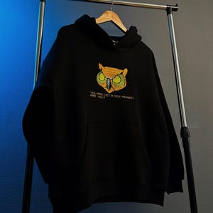 Pu&ograve; includere: Felpa nera con tasca frontale e cappuccio, appesa a un supporto metallico. La felpa presenta un design di gufo ricamato in giallo, verde e arancione, con il testo "YOU ARE NOT A NICE PERSON, ARE YOU?" sotto.