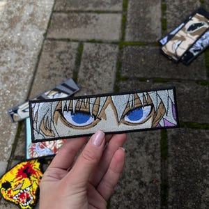 May include: Embroidered patch featuring anime-style eyes with blue irises, set against a white and tan background. The patch is rectangular with a black border. Several other patches with anime-style graphics are visible.