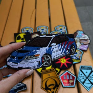 May include: Embroidered patch featuring a blue and white sports car with red accents. The patch is detailed with black outlines and a racing design. Other patches with various designs are visible in the background.
