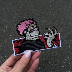May include: Embroidered patch depicting a character with pink, spiky hair, a sinister grin, and a clawed hand. The rectangular patch has a red and black background and a white border. The character is dressed in black.