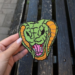 May include: A green and orange embroidered patch featuring a cobra with its mouth open. The patch has a black outline and is held by a hand. The cobra's mouth is pink and its eyes are black.