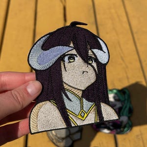 May include: An embroidered patch featuring an anime-style character with long dark purple hair, pale skin, and large yellow eyes. The character has curved light blue horns and wears a white and gold garment with a high collar and a diamond-shaped accent.