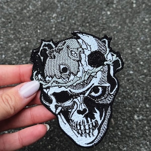 Könnte beinhalten: Gestickter Aufnäher mit einem detaillierten Totenkopf-Design in Schwarz und Hellblau. Der Schädel weist komplizierte Details auf, darunter Zähne, Augenhöhlen und ein kronenartiges Element. Der Aufnäher ist für Kleidung oder Accessoires.