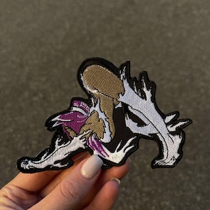 Puede incluir: Parche bordado con un diseño de criatura estilizada en negro, blanco, morado y marrón. El parche tiene un contorno detallado y es adecuado para ropa o accesorios.