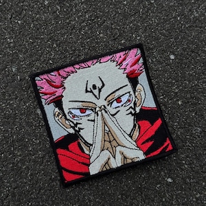 Puede incluir: Parche bordado cuadrado que representa un personaje de anime con cabello rosado y ojos rojos. El personaje se muestra con las manos juntas cerca de la cara, vistiendo un atuendo rojo. El parche tiene un borde negro.