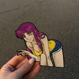 Könnte beinhalten: Gestickter Aufnäher mit einem Anime-Charakter mit lila Haaren, einem gelben Oberteil und Jeansshorts. Der Charakter zwinkert und gestikuliert. Der Aufnäher wird vor einem grauen Hintergrund gehalten.