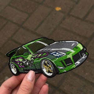 以下が含まれることがあります： 緑色のスポーツカーの刺繍パッチ。グレーのボンネットと黒いスポイラーが付いています。車体には紫色の炎と金色のリムを持つドラゴンのグラフィックが描かれています。パッチは手で持たれています。