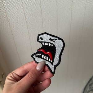 Könnte beinhalten: Gestickter Aufnäher mit einem schreienden Gesicht mit weit geöffnetem Mund. Das Design verwendet einen grauen Umriss, einen schwarzen Innenraum und einen roten Mund. Der Aufnäher wird vor einem neutralen Hintergrund gehalten.