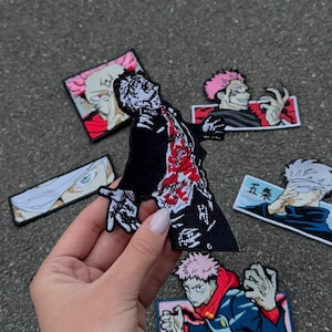 Può includere: Una collezione di toppe ricamate con personaggi di anime. Le toppe raffigurano vari personaggi in pose dinamiche, con un design nero e rosso di spicco. Le toppe sono su una superficie grigia.