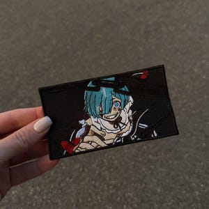May include: Black rectangular patch featuring an anime character with blue hair and a sinister expression. The character is wearing a white outfit with red accents. The patch is embroidered with detailed stitching.