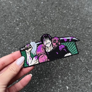 May include: Embroidered patch depicting two figures in a dynamic pose. One figure holds a weapon, the other wears a mask. The patch has a black background with purple and green accents. The patch is held by a hand.