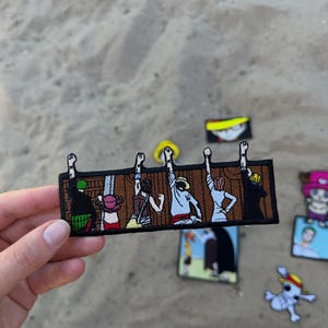 May include: A rectangular embroidered patch featuring six cartoon characters standing in a row with their backs turned, each raising one arm with a black mark on their wrist. The characters are set against a brown wooden fence background.