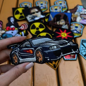 May include: Embroidered patch of a black sports car with orange and red flames. The patch is held in front of a collection of other patches featuring various designs, including a nuclear symbol and cartoon characters.