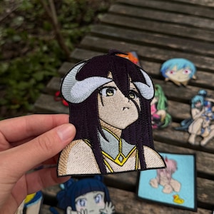May include: An embroidered patch featuring a stylized anime character with long dark purple hair, large light-colored horns, and yellow eyes. The character wears a light-colored garment with gold trim. The patch has a distinct dark border.