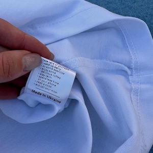 Pu&ograve; includere: Primo piano di una maglietta bianca con un'etichetta. L'etichetta contiene consigli per la cura di s&eacute; e la scritta "Made in Ukraine".