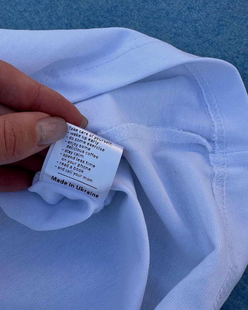 Pu&ograve; includere: Primo piano di una maglietta bianca con un'etichetta. L'etichetta contiene consigli per la cura di s&eacute; e la scritta "Made in Ukraine".