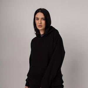 Pu&ograve; includere: Felpa con cappuccio nera e pantaloni abbinati. La felpa ha un cappuccio con coulisse e una tasca a marsupio. I pantaloni sono di vestibilit&agrave; ampia. L'outfit &egrave; realizzato in un materiale morbido e confortevole. La modella indossa una maglietta viola.