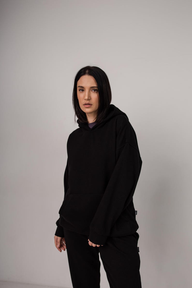 Pu&ograve; includere: Felpa con cappuccio nera e pantaloni abbinati. La felpa ha un cappuccio con coulisse e una tasca a marsupio. I pantaloni sono di vestibilit&agrave; ampia. L'outfit &egrave; realizzato in un materiale morbido e confortevole. La modella indossa una maglietta viola.