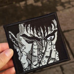 May include: A square embroidered patch featuring a black and white graphic illustration of a character with short dark hair, intense eyes, and fingers positioned over the face. The design is rendered in high-contrast thread on a black background.