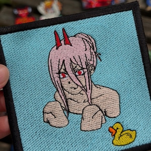 May include: A square embroidered patch featuring a character with light pink hair, two red horns, and red eyes against a light blue background. The character is depicted from the chest up, and a small yellow rubber duck is positioned in the bottom right corner.