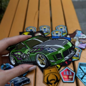 Peut inclure: Écusson de voiture brodé vert avec un capot gris et des roues dorées. Le patch présente un design détaillé d'une voiture de sport avec un motif de dragon sur le côté. D'autres écussons sont visibles en arrière-plan.