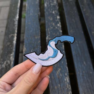 May include: Embroidered patch with a wave design in blue and white. The patch has a black border. The wave design is a popular motif for clothing and accessories. The patch is held against a wooden bench.