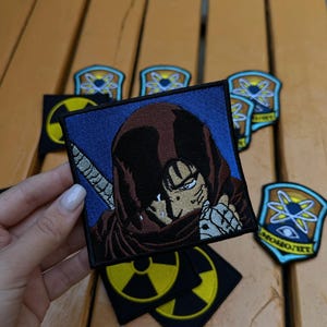 May include: Embroidered patches featuring anime-style characters and symbols. The main patch shows a character with a brown hood and a sword. Other patches include a yellow hazard symbol and a blue and yellow emblem.