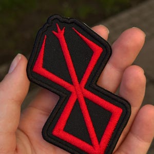 Może przedstawiać: Czarna haftowana naszywka z wyrazistym czerwonym stylizowanym symbolem. Wzór składa się z centralnej pionowej linii z trzema ząbkami u góry, przecinającej się z geometrycznym kształtem przypominającym kwadrat z jednym otwartym bokiem.