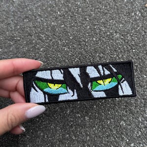 May include: A black rectangular patch featuring a stylized illustration of two eyes. The eyes have green irises with yellow highlights and blue accents. The patch is bordered by white and black details.