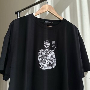 Op de afbeelding: Zwart t-shirt met een witte afbeelding van een persoon met een zwaard, met de tekst "THINK ONLY ABOUT YOUR ART". Het t-shirt hangt aan een houten hanger.