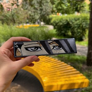 以下が含まれることがあります： 額に黒い髪の毛がかかった、鋭い眼差しの目元が描かれた長方形の刺繍ワッペン。黒、グレー、ベージュの糸で構成されており、周囲は太い黒の縁取りで囲まれています。