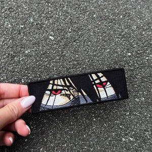 May include: Black rectangular patch with an anime-style eye illustration, featuring red irises and black hair. The patch is held against a textured grey surface. The eyes have a light skin tone.