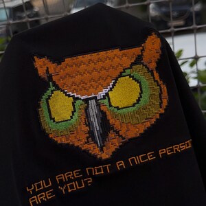 Pu&ograve; includere: Tessuto nero con un disegno di gufo ricamato nei toni dell'arancione, verde e giallo. Il testo "YOU ARE NOT A NICE PERSON ARE YOU?" &egrave; ricamato in arancione sotto il gufo.