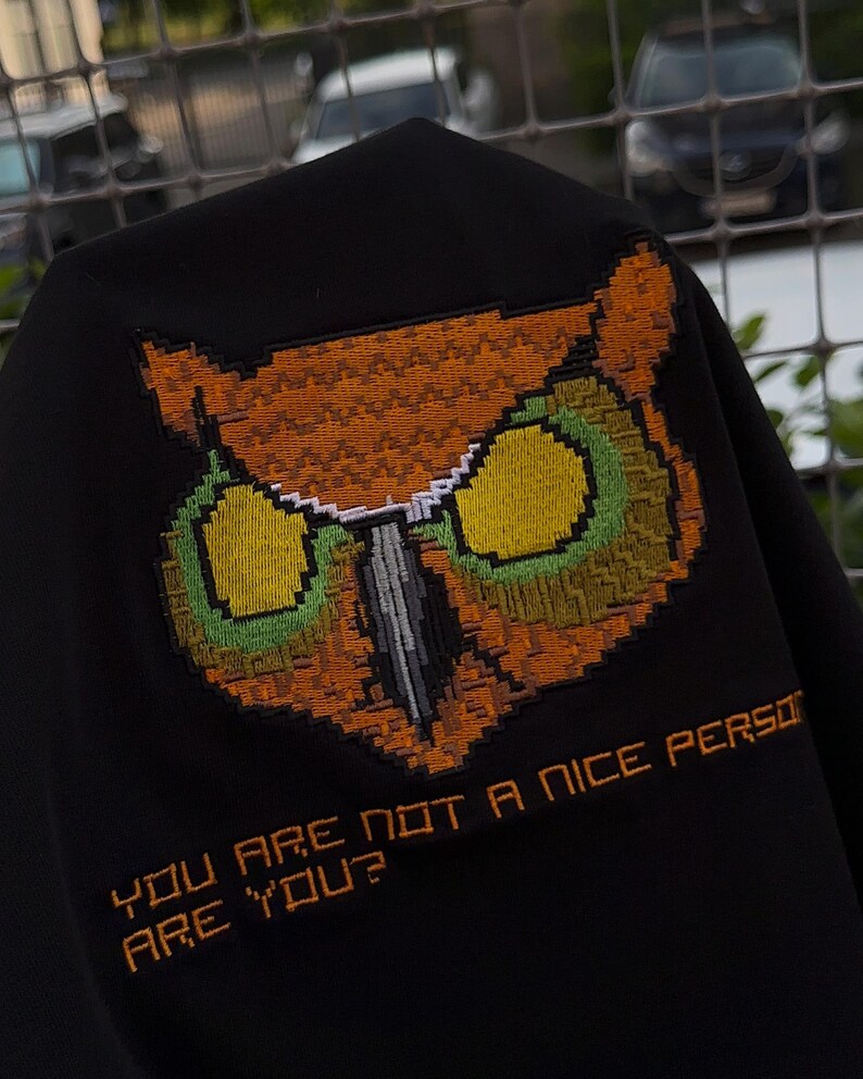 Pu&ograve; includere: Tessuto nero con un disegno di gufo ricamato nei toni dell'arancione, verde e giallo. Il testo "YOU ARE NOT A NICE PERSON ARE YOU?" &egrave; ricamato in arancione sotto il gufo.