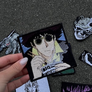 Peut inclure: Plusieurs écussons brodés carrés avec des motifs de style anime. L'un d'eux représente un personnage avec des lunettes rondes mangeant des nouilles, avec le texte "BIG" visible. D'autres écussons présentent des crânes et des personnages d'anime.