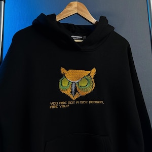 Pu&ograve; includere: Felpa con cappuccio nera con un design a gufo pixelato nei toni del giallo, verde e arancione. Il testo "YOU ARE NOT A NICE PERSON, ARE YOU?" &egrave; stampato sotto il gufo. La felpa &egrave; appesa a una gruccia di legno.