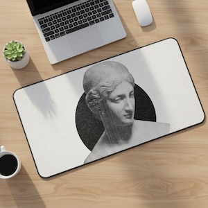 May include: A rectangular mouse pad featuring a black and white illustration of a classical bust against a black circle. The mouse pad has a black border and is on a wooden desk with a laptop, mouse, and a small potted plant.