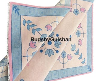 May include: A rolled-up area rug with a floral design. The rug features a light beige background with blue and pink floral patterns and a blue border. The text "RugsbyGulshan" is visible on the rug.