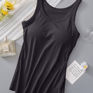Könnte beinhalten: Ein dunkelgraues Tanktop mit Rundhalsausschnitt und breiten Trägern. Das Oberteil ist aus einem weichen, glatten Stoff gefertigt. Eine kleine Karte mit Text und einer Gänseblümchen liegt neben dem Tanktop. Ein Parfümflakon und gelbe Blumen sind ebenfalls im Bild.