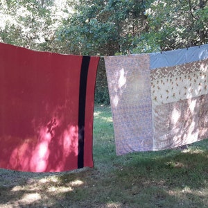 May include: Two blankets hanging on a clothesline outdoors. The blanket on the left is red with black stripes. The blanket on the right is a patchwork of different colors and patterns.