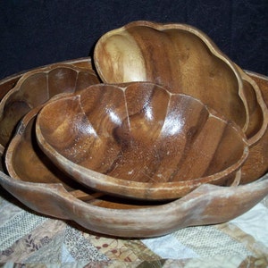 Peut inclure: Un ensemble de bols en bois de diff&eacute;rentes tailles, embo&icirc;t&eacute;s les uns dans les autres. Les bols ont une finition en grain de bois naturel et un bord festonn&eacute;. Les bols sont dispos&eacute;s sur un tissu &agrave; motifs.