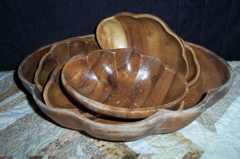 Peut inclure: Un ensemble de bols en bois de diff&eacute;rentes tailles, embo&icirc;t&eacute;s les uns dans les autres. Les bols ont une finition en grain de bois naturel et un bord festonn&eacute;. Les bols sont dispos&eacute;s sur un tissu &agrave; motifs.