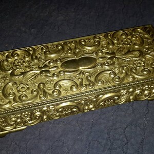 May include: A gold-colored metal trinket box with an ornate, embossed floral design. The box has a rectangular shape and a hinged lid.