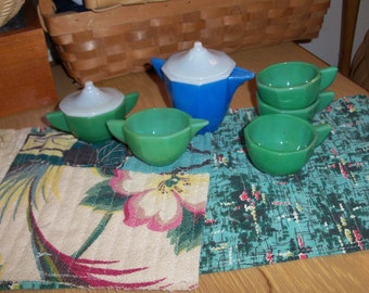 May include: A blue and white teapot with a lid and a handle, surrounded by four green teacups with white rims. The teacups are on a green and white patterned tablecloth.